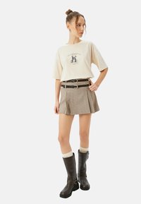 Camiseta gráfica beige con diseño de panda, combinada con una falda de cuadros marrón. Botas altas marrón con tiras negras y un acabado texturizado.