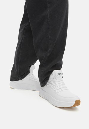 Scarpe da ginnastica bianche con superficie texturizzata, dotate di lacci, suola spessa e dettagli in gomma color caramello, abbinate a jeans scuri e larghi.