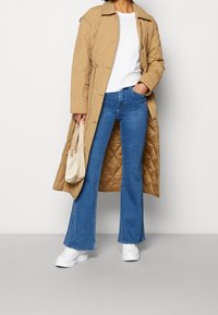 Quiltad beige lång kappa, vit t-shirt, utsvängda blå jeans och vita sneakers. Har en knappstängning och sidofickor.