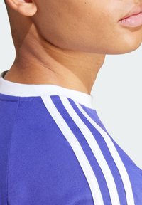 Fioletowa koszulka sportowa z białym okrągłym dekoltem i trzema białymi paskami na ramionach, o gładkiej teksturze i dopasowanym kroju.