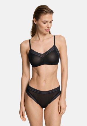 Vrouw die een zwarte doorschijnende beha en bijpassende hoge taille slip draagt met kanten rand, staand en naar beneden kijkend tegen een effen achtergrond.