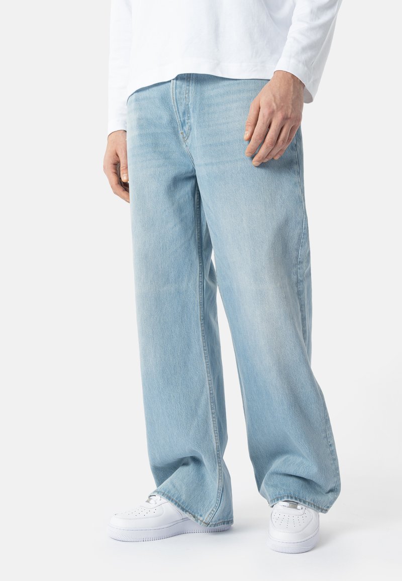 Personne portant un jean large bleu clair, une chemise blanche à manches longues et des baskets blanches, debout devant un fond uni.