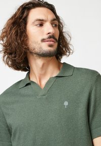 Grün gestricktes Polohemd mit V-Ausschnitt und kurzen Ärmeln. Verfügt über ein dezentes Baumlogo auf der linken Brust. Glatte Textur.