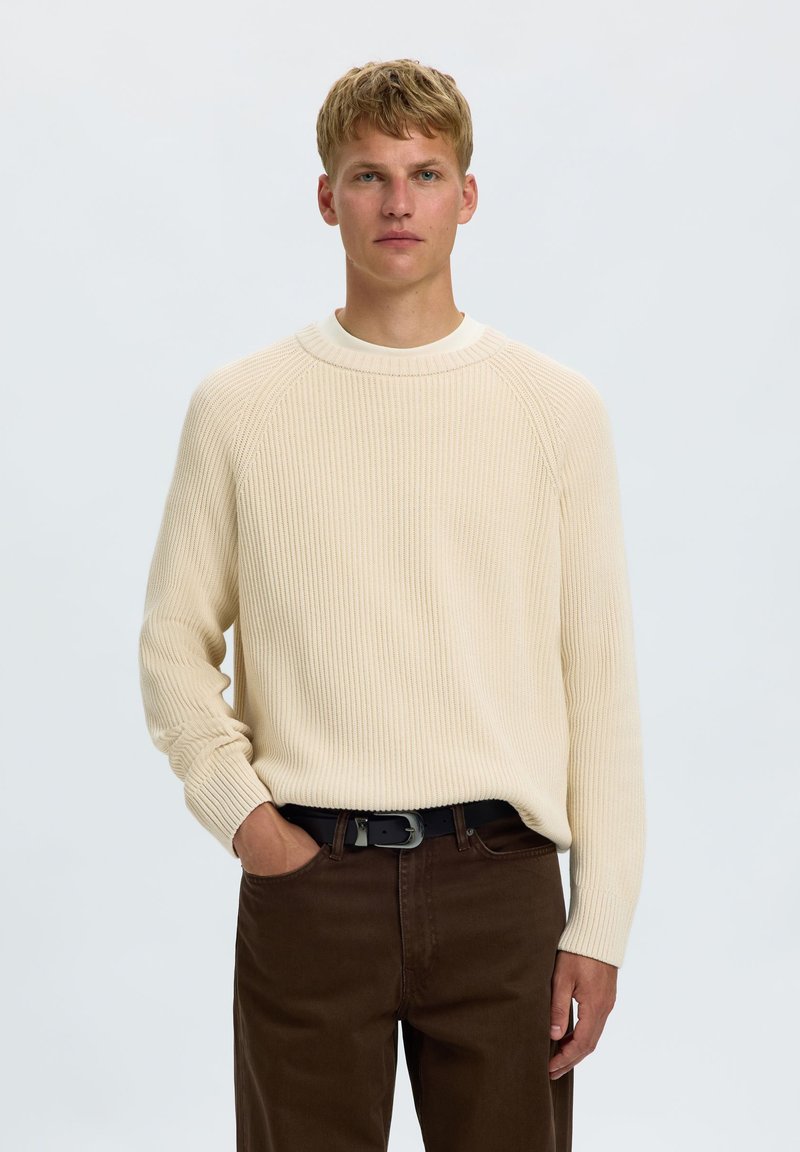 Jeune homme aux cheveux blonds courts, portant un pull côtelé crème et un pantalon marron foncé, debout avec une main dans la poche devant un fond uni.