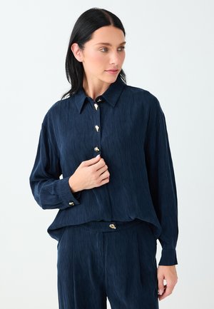 Femme aux cheveux foncés portant une chemise à manches longues bleu marine texturée et un pantalon assorti avec des boutons dorés uniques, posant devant un fond uni.