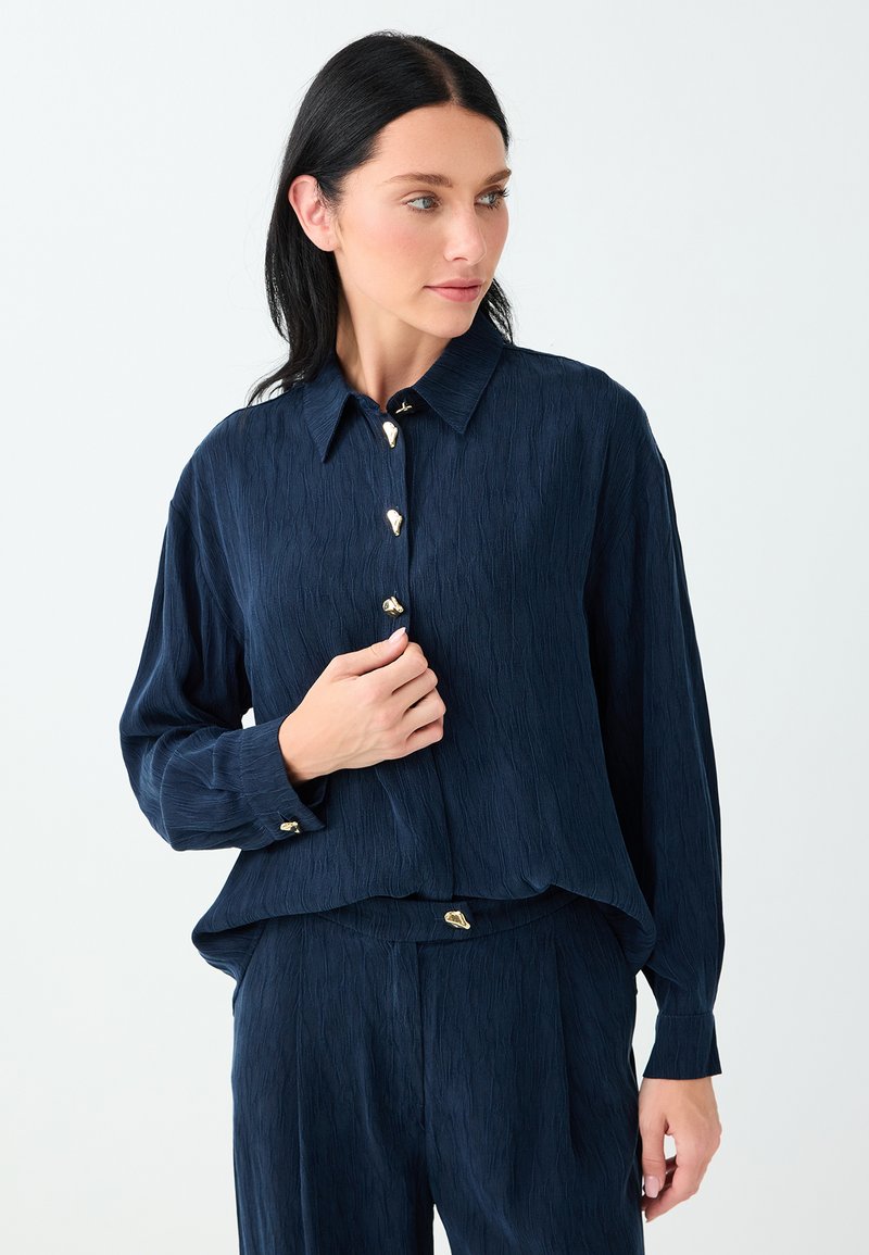 Femme aux cheveux foncés portant une chemise à manches longues bleu marine texturée et un pantalon assorti avec des boutons dorés uniques, posant devant un fond uni.