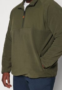 Uomo che indossa un pullover in pile verde oliva con mezzo zip e tasca laterale, abbinato a pantaloni blu scuro su uno sfondo semplice.