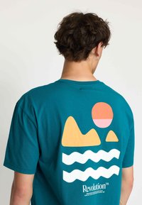 Teal katoenen t-shirt met een grafisch ontwerp van een zon, bergen en golven in oranje en wit op de achterkant, met logo-tekst onderaan.