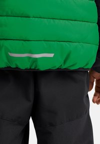 Grüne Pufferjacke mit horizontaler Steppung und einem reflektierenden Streifen am unteren Rand; kombiniert mit schwarzen, glatttexturierten Hosen.