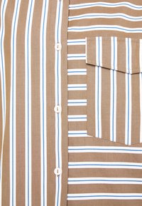 Gros plan sur une chemise beige avec des rayures blanches et bleues verticales et horizontales, des boutons blancs et une poche avant avec rabat.
