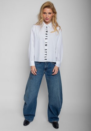 Blondynka w białej koszuli z napisem "ALWAYS IN STYLE" i szerokich niebieskich jeansach stojąca na jednolitym jasnym tle.