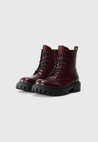 Par de botas de cuero burdeos con cordones, suelas de goma negra gruesa y cremalleras laterales sobre un fondo blanco.