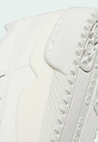 Lähikuva valkoisen Adidas-tennin kantapäästä, jossa on teksturoitu pohja, ompelu ja kohokuvioitu Adidas-logo kumipohjassa.