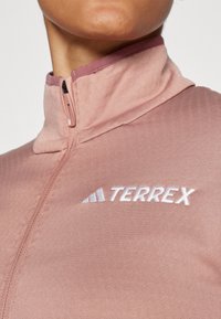 Svijetloružičasta jakna na zip s visokim ovratnikom, koja sadrži bijelu izvezenu "TERREX" oznaku na prsima, glatke teksture i jednostavnog dizajna.