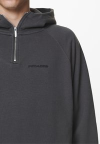 Grå zip-up hoodie med hög krage, ribbad textur, kontrasterande svart logotyp på bröstet och silverfärgad dragkedjehårdvara.