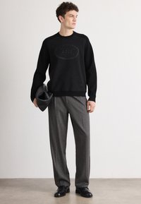 Maglione nero con logo testurizzato, abbinato a pantaloni grigi a quadri. Il modello tiene una pochette nera; l'ambientazione è minimalista e neutra.