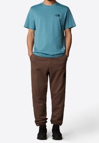 Conjunto casual masculino que incluye una camiseta de manga corta en color azul verdoso y pantalones de chándal marrones con bolsillos y un parche con logo, acompañados de zapatos negros.