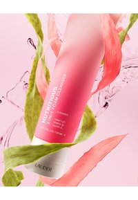 Skumrensare i en rosa tub med ett vitt lock. Texten lyder "Nutrient 2-i-1 Skumrensare." Omgiven av gröna och rosa löv, med vattendroppar.