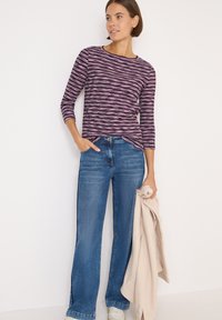 Maglione a maniche lunghe a righe magenta e nere abbinato a jeans blu a gamba larga. Il modello tiene un maglione beige e indossa sneakers bianche.