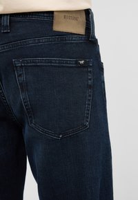 Ciemnoniebieskie dżinsy o gładkiej teksturze, z brązową skórzaną naszywką marki na tylnej części pasa oraz małą metką na tylniej kieszeni.