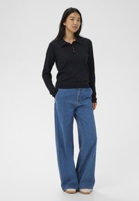 Egy nő áll, kezét zsebre dugva, fekete, hosszú ujjú póló pulóverben, széles szárú kék farmerban és fehér cipőben egy egyszerű háttér előtt.