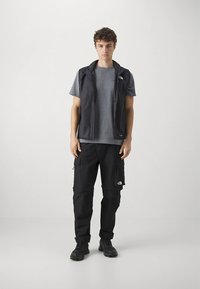 Mustast lõhnav vest halli T-särgi peal, mustade cargo-pükstega. Vestil on lukuga ja vasakul rinnal logoga detail.