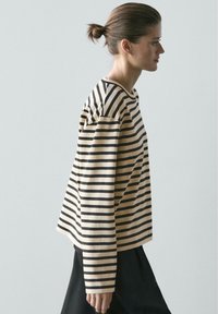 Frau mit braunen Haaren zu einem Dutt gebunden, trägt ein langärmeliges, beige-schwarz gestreiftes Shirt und schwarze Hosen, blickt nach rechts vor einem schlichten Hintergrund.