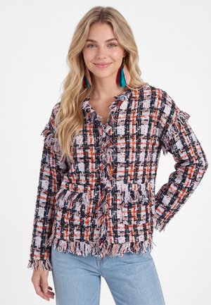 Giovane donna che indossa una giacca scozzese multicolore con frange, grandi orecchini di piume e jeans azzurri, sorridente con la mano sui fianchi.
