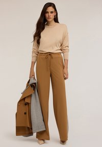 Maglione beige con spalle abbassate, pantaloni marroni con vita regolabile, e cappotto grigio appoggiato sull'avambraccio. Tessuto liscio e vestibilità sartoriale.