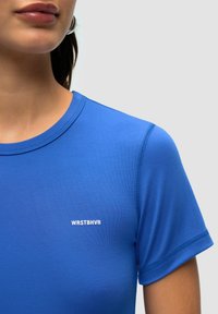 Niebieska koszulka sportowa wykonana z gładkiego materiału, z krótkimi rękawami i okrągłym dekoltem. Z przodu widoczny charakterystyczny biały logo "WRSTBHV".