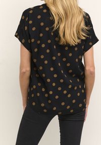 Mujer con cabello largo y rubio, vestida con una camiseta negra con lunares marrones y pantalones negros, vista desde la espalda contra un fondo liso.