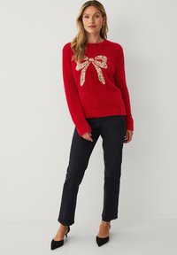 Rode, pluizige trui met een groot, gouden glitterstrikontwerp op de voorkant, gecombineerd met donkere jeans en zwarte hakken. Eenvoudige, ronde halslijn.