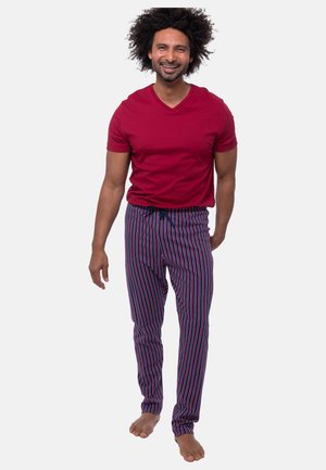 Mężczyzna o kręconych włosach, uśmiechający się, ubrany w czerwoną koszulkę z krótkim rękawem i niebieskie pasiasty spodnie piżamowe, stoi boso na białym tle.