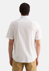 Camisa blanca de manga corta con una textura suave, cuello puntiagudo y dobladillo curvado. La parte de atrás presenta un discreto detalle de costura. Combinada con pantalones color terracota.