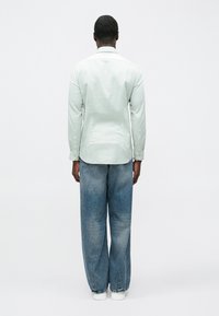 Šviesiai žalia ilgarankovė marškinėliai su sagomis, su viengubu rankogaliu, derinami su šviesiai mėlynais plačiais džinsais ir baltais sportbačiais, matomi iš nugaros.