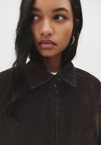 Rjava jeans jakna z ovratnikom, z zadrgo in modrimi šivi. Model ima dolge temne lase in mehko kožo.