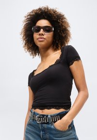 Γυναίκα με σγουρά μαλλιά που φοράει μαύρα γυαλιά ηλίου, μαύρο τοπ με δαντέλα, ζώνη με στέκες και μπλε τζιν, με το ένα χέρι στην τσέπη.