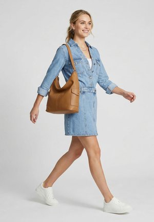 Woman walking wearing a light blue denim dress and white sneakers, carrying a large tan leather shoulder bag.