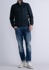 Mężczyzna w czarnej kurtce na zamek, w niebieskich wyblakłych dżinsach z mankietami i białych trampkach stoi na jednolitym jasnym tle.