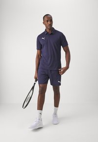 Marinefarbendes Polohemd und Shorts aus leichtem Material, mit einem weißen Logo. Das Model hält einen Tennisschläger und trägt weiße Sportschuhe und Socken.