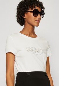 Camiseta blanca de algodón con mangas cortas, que presenta un logo de "GUESS" decorado con strass plateados. Combinada con pantalones negros de cintura alta.