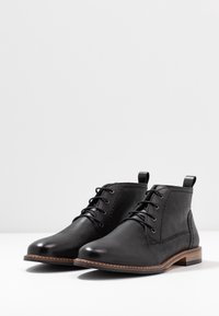 Botas de tobillo de cuero negro con punta redonda, cordones, costuras en contraste y un tacón cuadrado marrón. Incluyen lengüetas para facilitar el calzado.