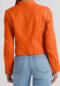 Giacca di pelle arancione dal design corto, con colletto piatto, maniche lunghe e polsini con cerniera, abbinata a jeans in denim azzurro.