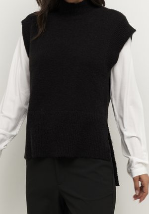 Vrouw in een mouwloze zwarte gebreide vest over een witte lange mouwen blouse en zwarte broek, staand tegen een effen lichte achtergrond.