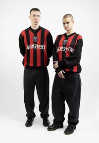 Des pulls rayés noirs et rouges avec le logo "Wasted", portés sur des jeans noirs amples et des bottes noires, présentés sur deux silhouettes debout.
