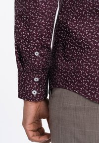 Bordeaux shirt met een klein wit bloemenpatroon, voorzien van knoopsluiting bij de manchetten en een zachte stof. Grijs geruite broek is gedeeltelijk zichtbaar.