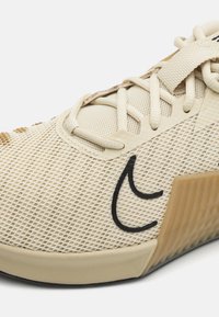 Beige athletic shoe with mesh upper, lace closure, black logo detail, and textured accents on the side and sole.