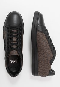 Svarta sneakers med en slät lädertå och snörning, bruna mönstrade textilsidor och en platt gummisula. Diskret varumärke synligt.