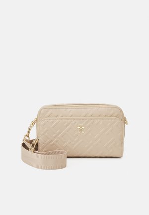 Beige quiltet crossbody-taske med logo og hardware i guldfarvet metal, frontlynlåslomme og justerbar stofrem på hvid baggrund.