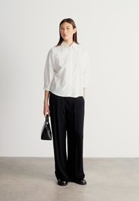 Λευκό πουκάμισο με κουμπιά και άνετη γραμμή, μαύρο παντελόνι με φαρδιά μπατζάκια, μαύρη τσάντα και μαύρα παπούτσια σε ουδέτερο φόντο. Ομαλές υφές, μινιμαλιστικός σχεδιασμός.
